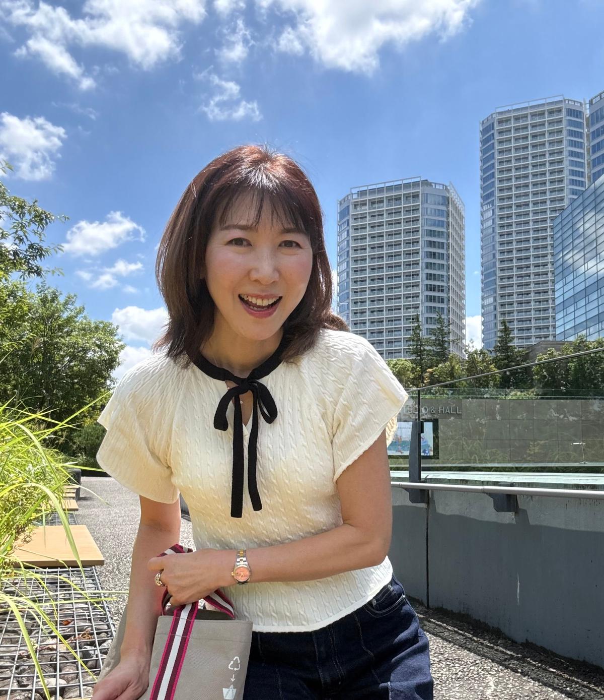 白いトップスにネイビーのワイドパンツを着用した女性が、屋外の歩道に立っている。背景には緑豊かな木々と高層ビルが見える。女性は笑顔で、赤いスカーフを巻いている。