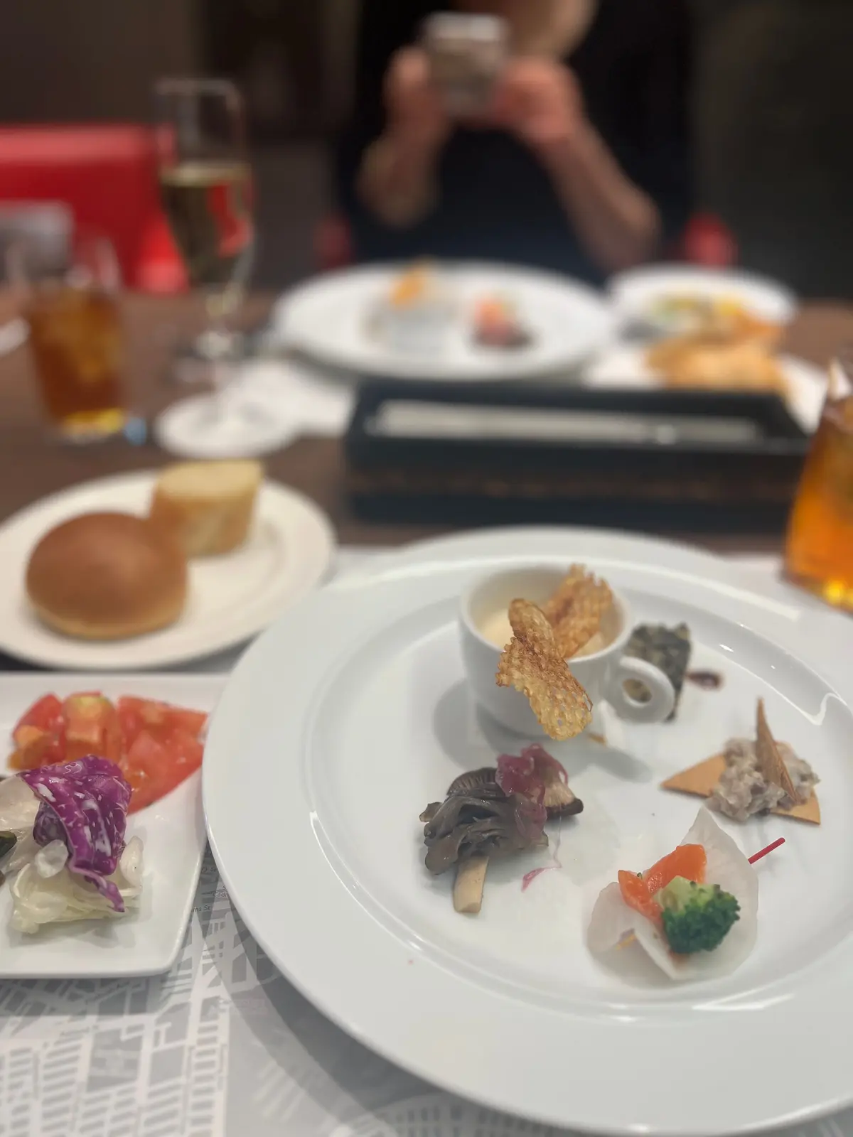 白磁の皿に、様々な種類のカナッペが盛り付けられている。前菜の盛り合わせには、魚介、野菜、パンなどが使われている。背景には、他の客のテーブルや飲み物が見える。