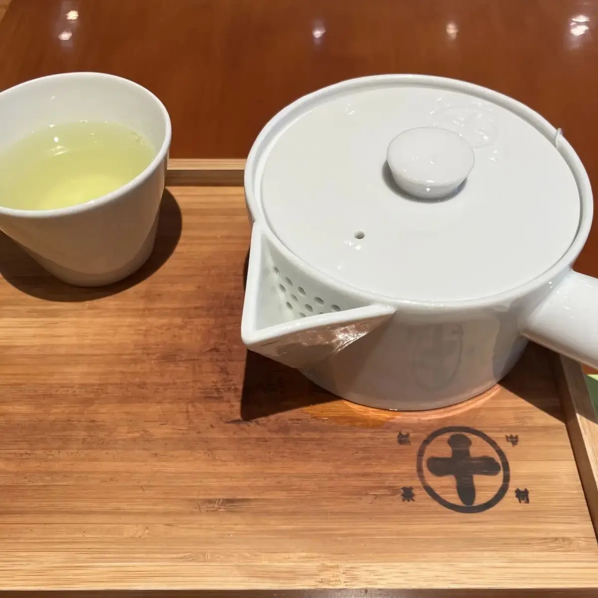 白磁製の茶器と湯呑み。茶器は丸みを帯びた形、蓋には取っ手が付く。湯呑みには淡い緑色の緑茶が入っている。木製のトレイに置かれ、中央には十字の模様がある。