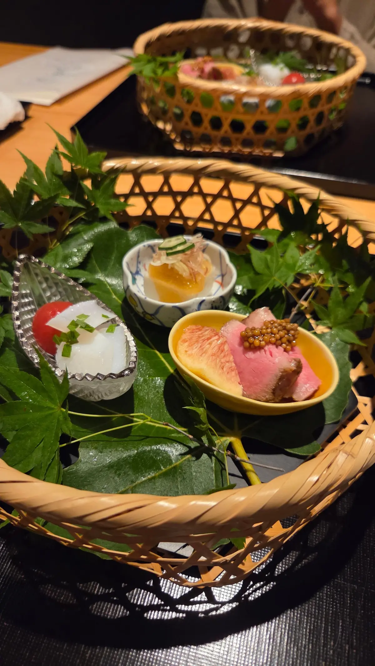 竹製の盛り合わせに、様々な日本料理が盛り付けられている。銀色の容器には刺身、小皿には肉とイクラ、ガラスの器にはゼリー状の料理が入っている。緑の葉が添えられ、上品な盛り付けが施されている。