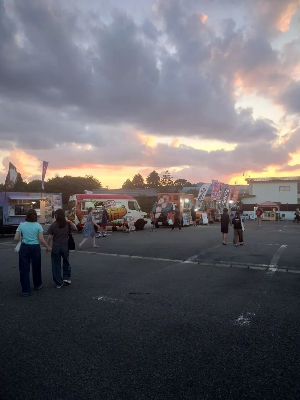 夕焼け空の下、複数のフードトラックが並ぶイベント会場。人々がトラックの間を歩き、食事を楽しんでいる様子。背景には樹木と建物が見える。