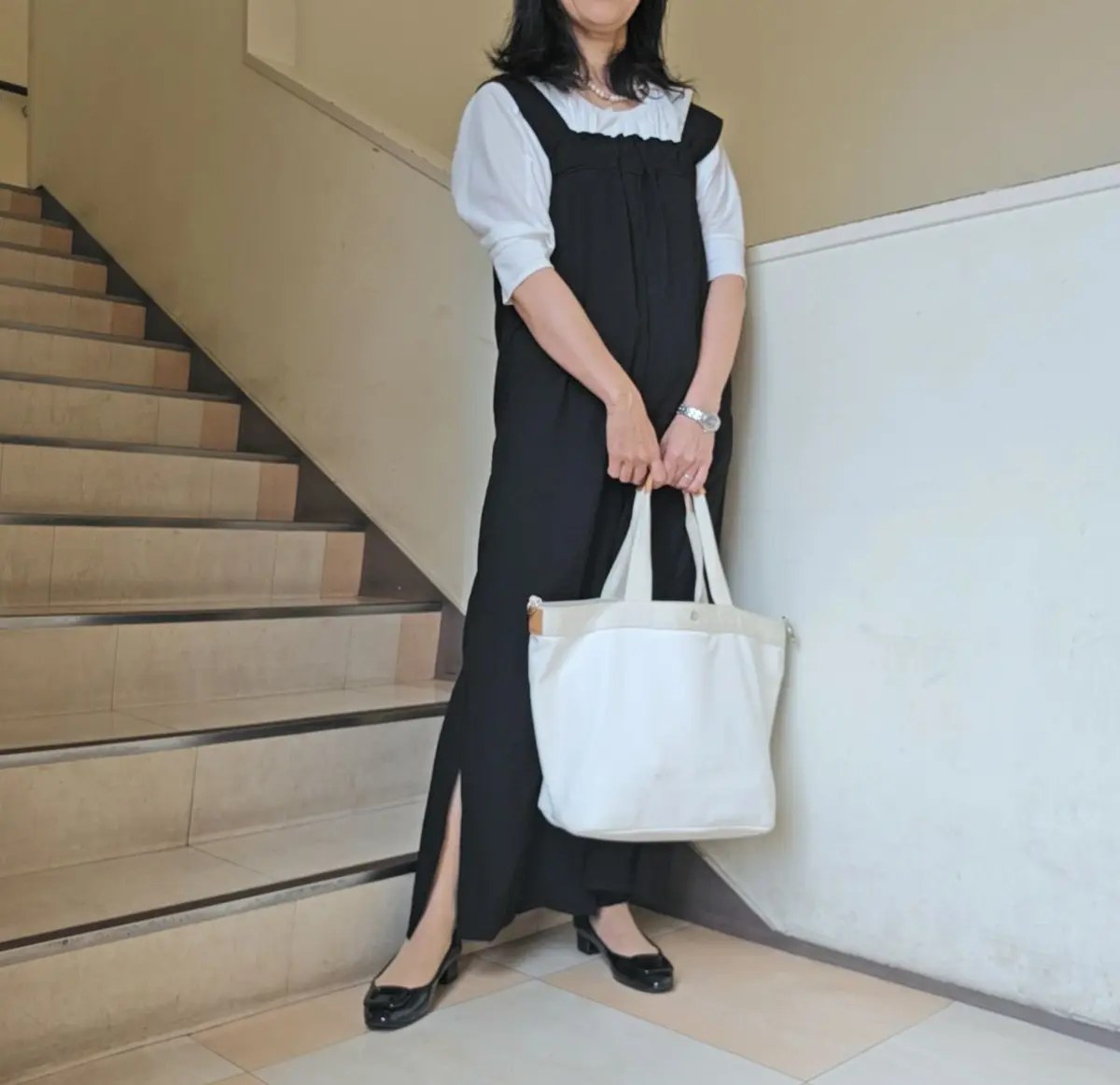 黒い無地のロングワンピースに白いブラウスを合わせた女性。右手に白いトートバッグを持ち、階段の横に立っている。黒いパンプスを履き、腕時計をしている。