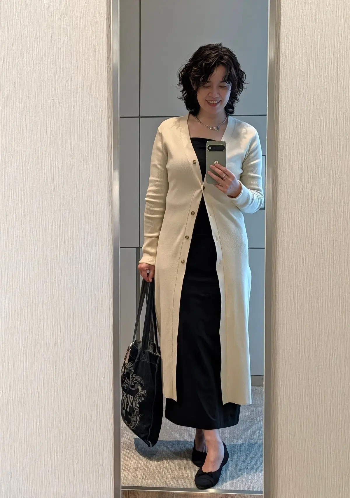 黒いワンピースにアイボリーのロングカーディガンを羽織った女性。黒いパンプスを履き、黒いバッグを持つ。笑顔でスマートフォンを操作している。