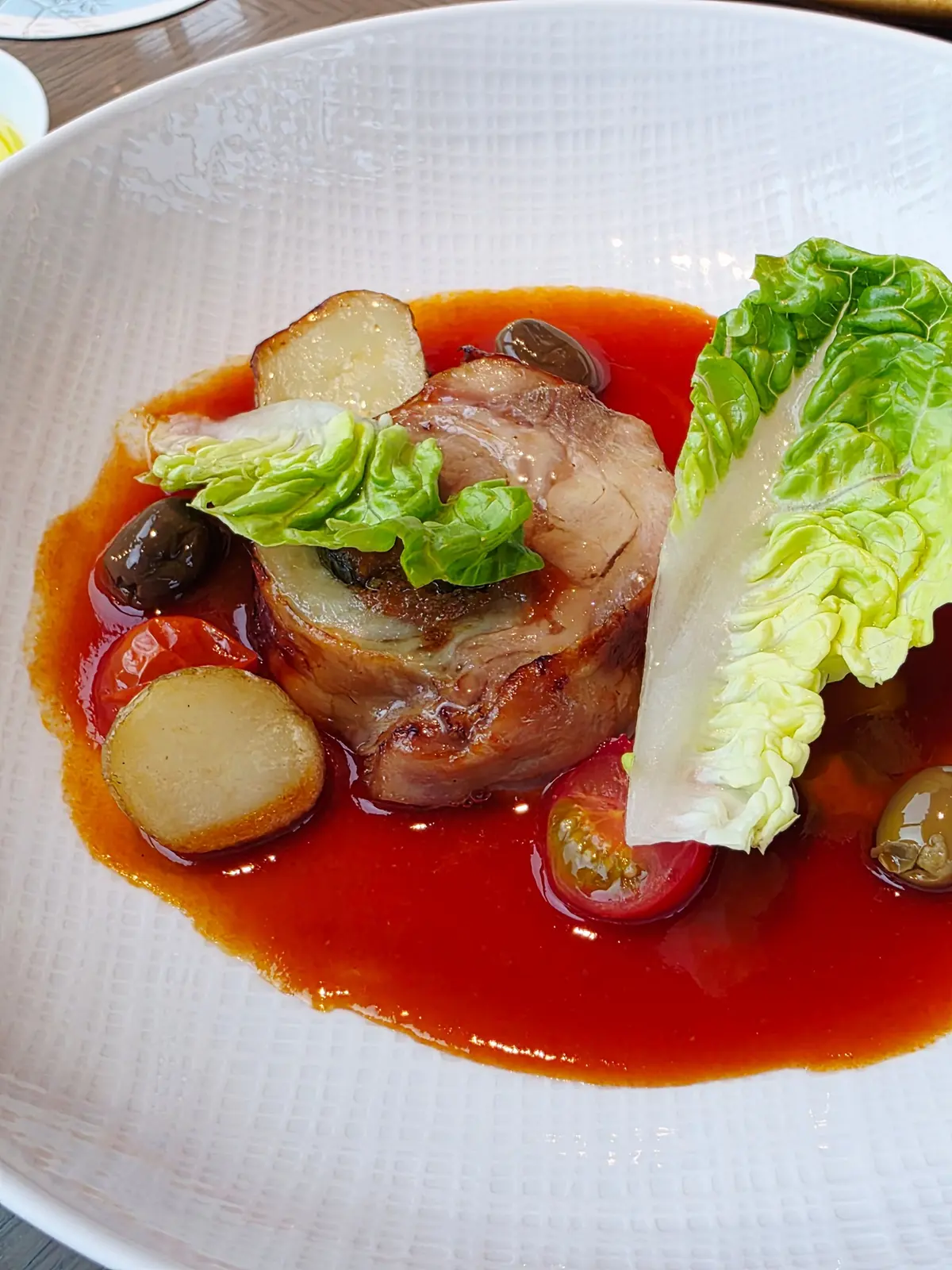 白い皿に盛り付けられた、豚肉料理。赤色のソースが広がり、その上に豚肉、薄切り大根、レタスが配置されている。黒い実（オリーブ？）とトマトも添えられている。