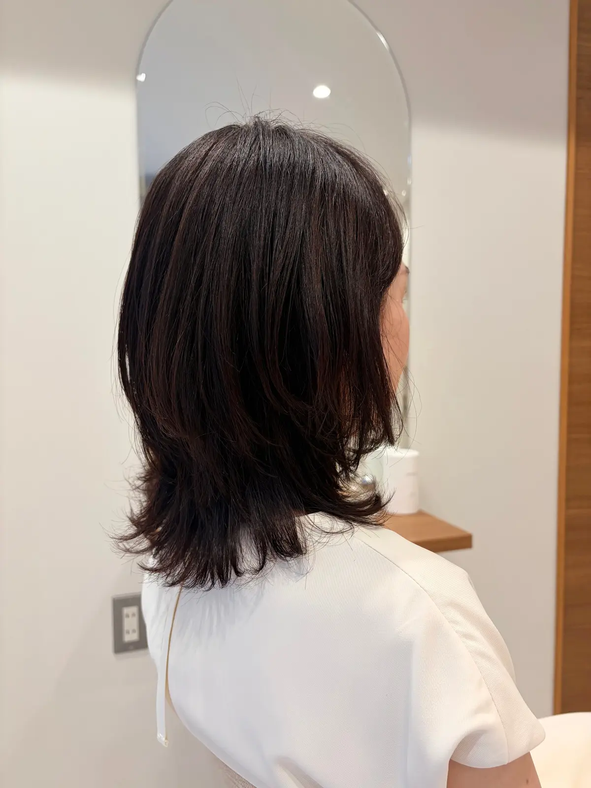 肩くらいの長さのダークブラウンのボブヘア。光沢があり、毛先が外側にカールしている。白いトップスを着た女性の背面からの写真。背景には鏡と木製の棚が見える。