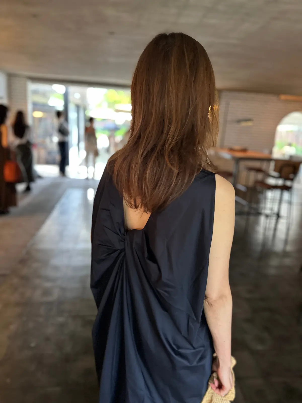 黒いワンピースを着た女性の後ろ姿。肩から背中にかけてドレープが施され、長い茶色の髪が肩にかかっている。背景には、窓から光が差し込むカフェのような空間。