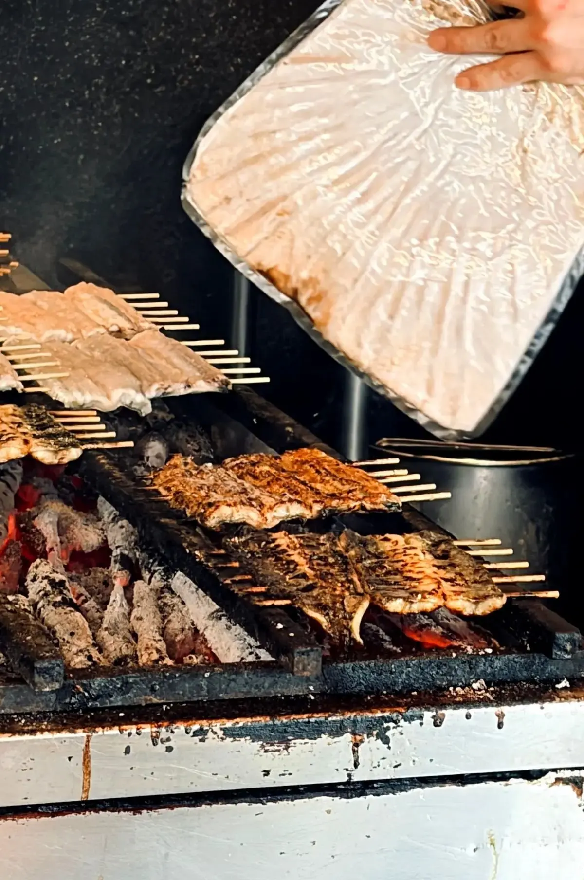 炭火のグリルで焼かれる肉。手で液体を注いでいる。グリルは金属製で、肉は串に刺され、焦げ目がついている。背景は暗く、煙が立ち込めている。