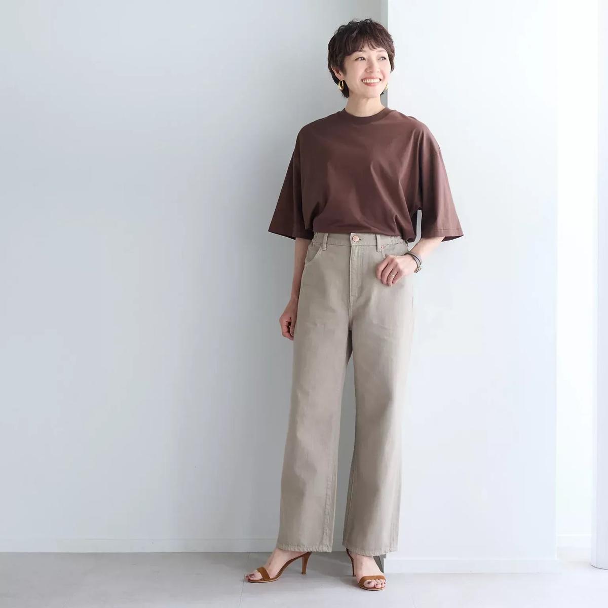 ブラウンのTシャツにベージュのワイドパンツを合わせた女性。右手を腰に当てて微笑み、シンプルな背景でポーズをとっている。