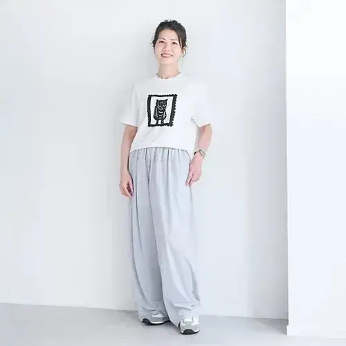 白い猫のイラストがプリントされた白いTシャツと、グレーのワイドパンツを着用した女性が立っている全身写真。背景は白。