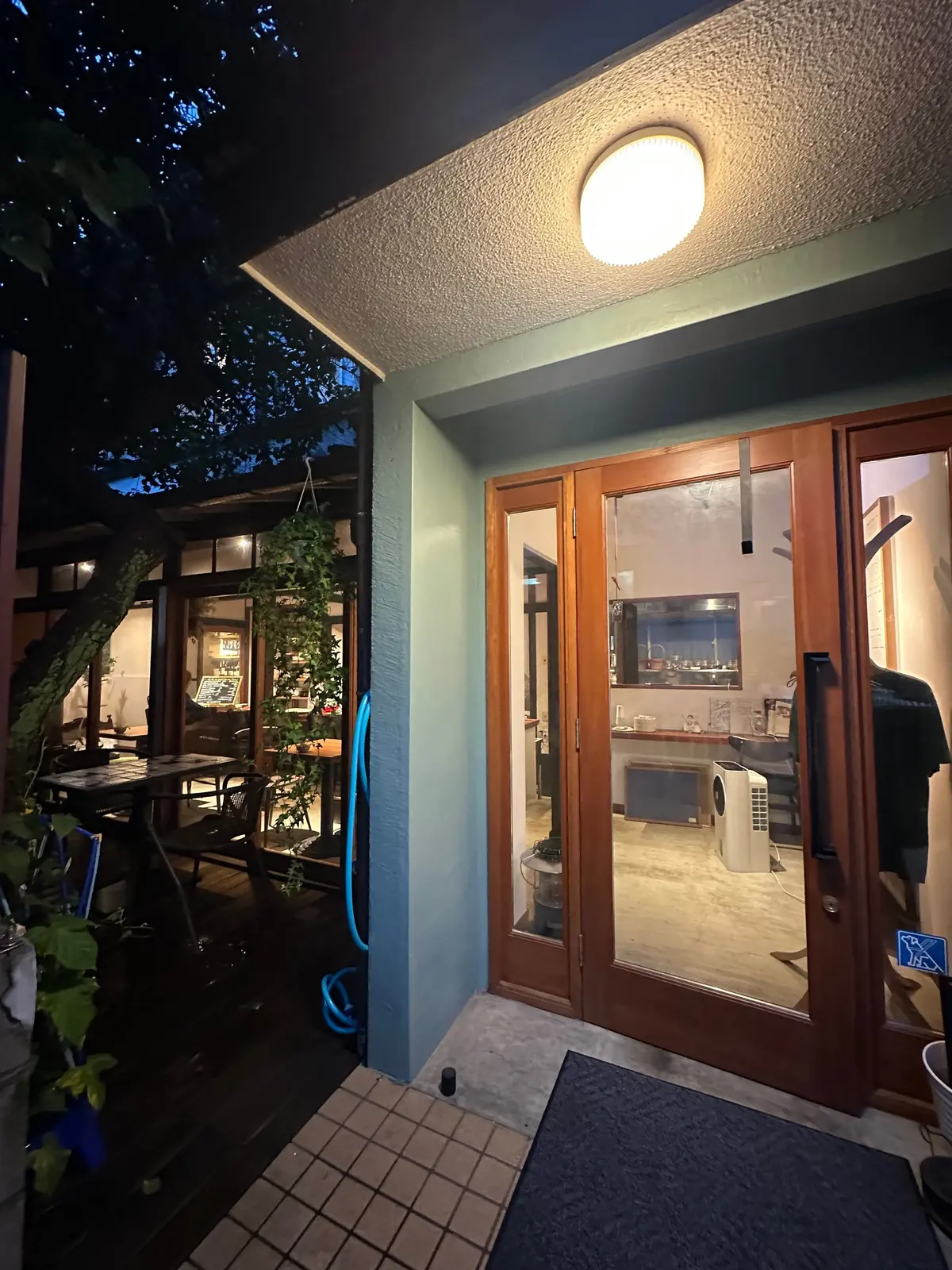 夜の屋外、木製の二重ドアと窓のある建物。右には丸い照明、左には木製のテーブルと椅子が並ぶパティオ。緑の植物が建物の壁を覆っている。