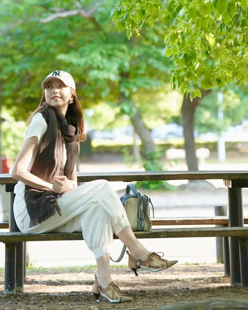 女性が木製ベンチに座り、木陰の公園でリラックスしている。白のキャップ、軽い服、スカーフを着用し、ベージュのハンドバッグをベンチに置いている。