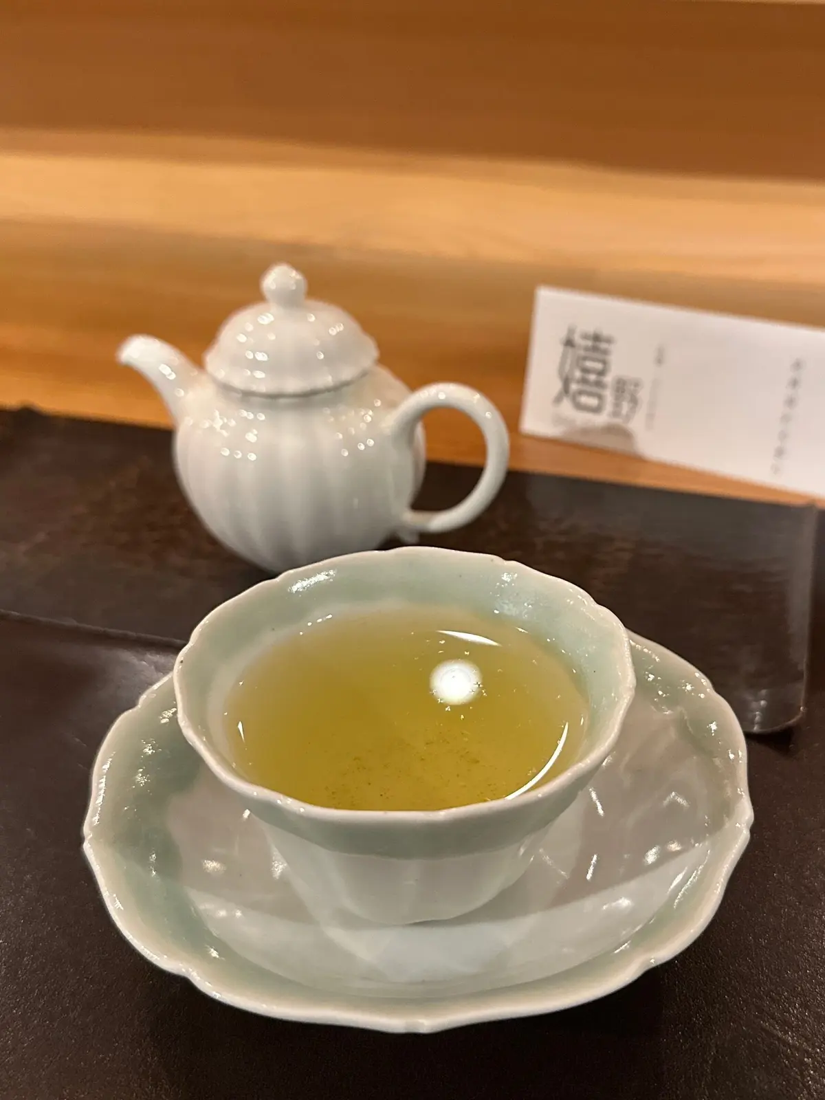 灰色のティーポットと、波型の縁が特徴的な灰色の湯呑み。湯呑みには淡い緑色の緑茶が入っており、光が反射している。背景には黒いマットと木製のテーブルがある。