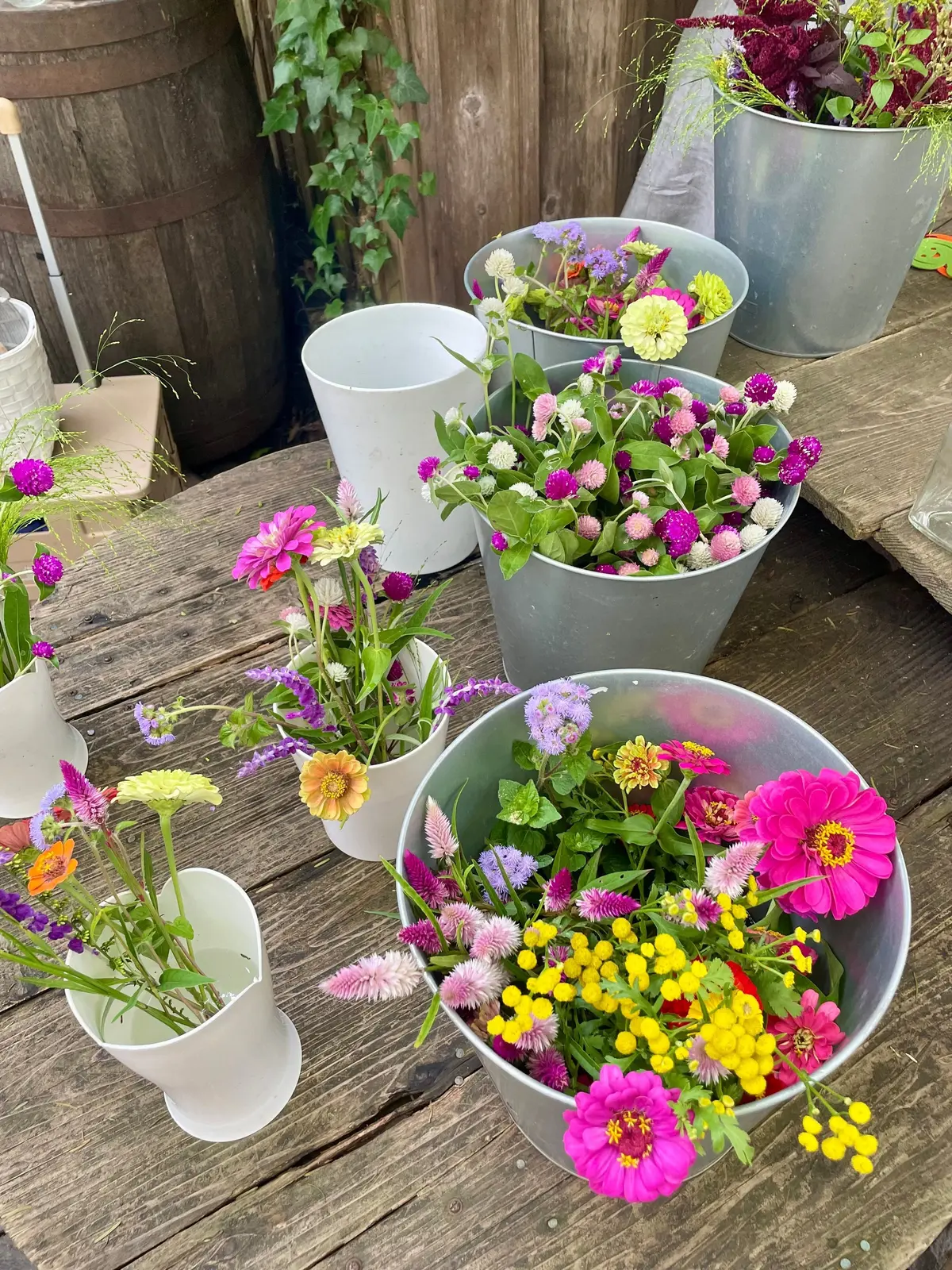 木製のテーブルに、様々な花を植えた白い植木鉢と銀色のバケツが並んでいる。紫、ピンク、黄色の花が咲き、背景には木製の樽と緑の植物が見える。