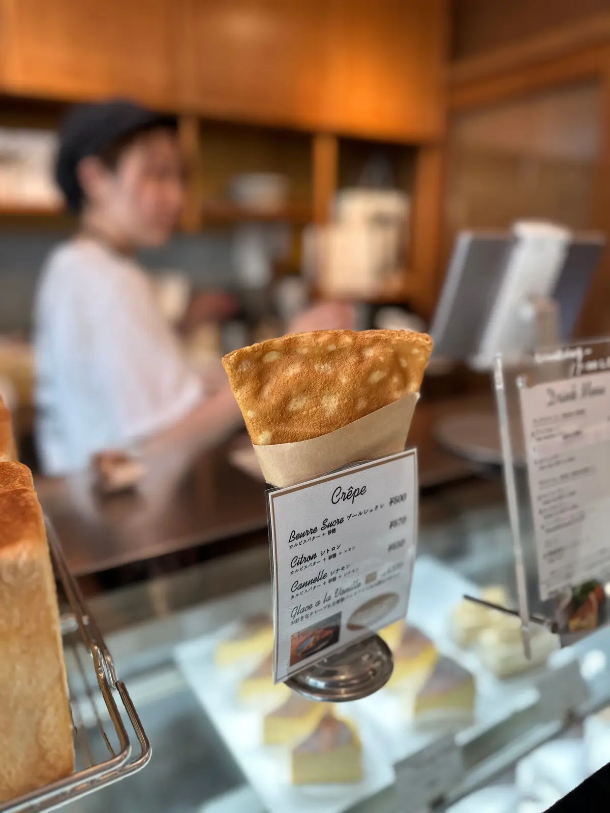 紙コーンに包まれたクレープが、メニューカードの前に置かれている。背景には、店員と思わしき人物が作業している。木目調の店内、自然光が差し込む様子。