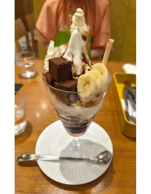 ガラスの器に入った、生チョコレート、バナナ、ホイップクリーム、ソフトクリーム、チョコレートソースを組み合わせたデザート。葉の飾りと、棒状のクッキーが添えられている。