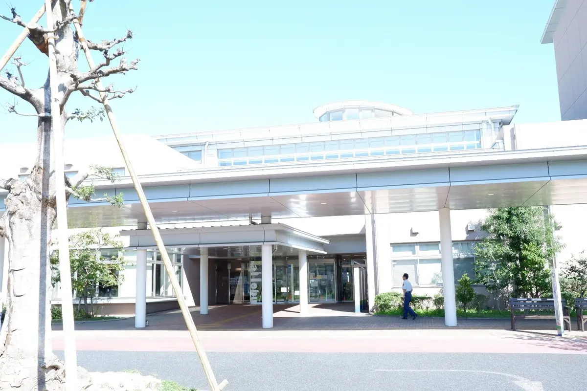 白い近代的な建物と、その入り口を覆う広い屋根。手前に木の枝が伸び、右には歩行者がいる。青空が背景に広がる。