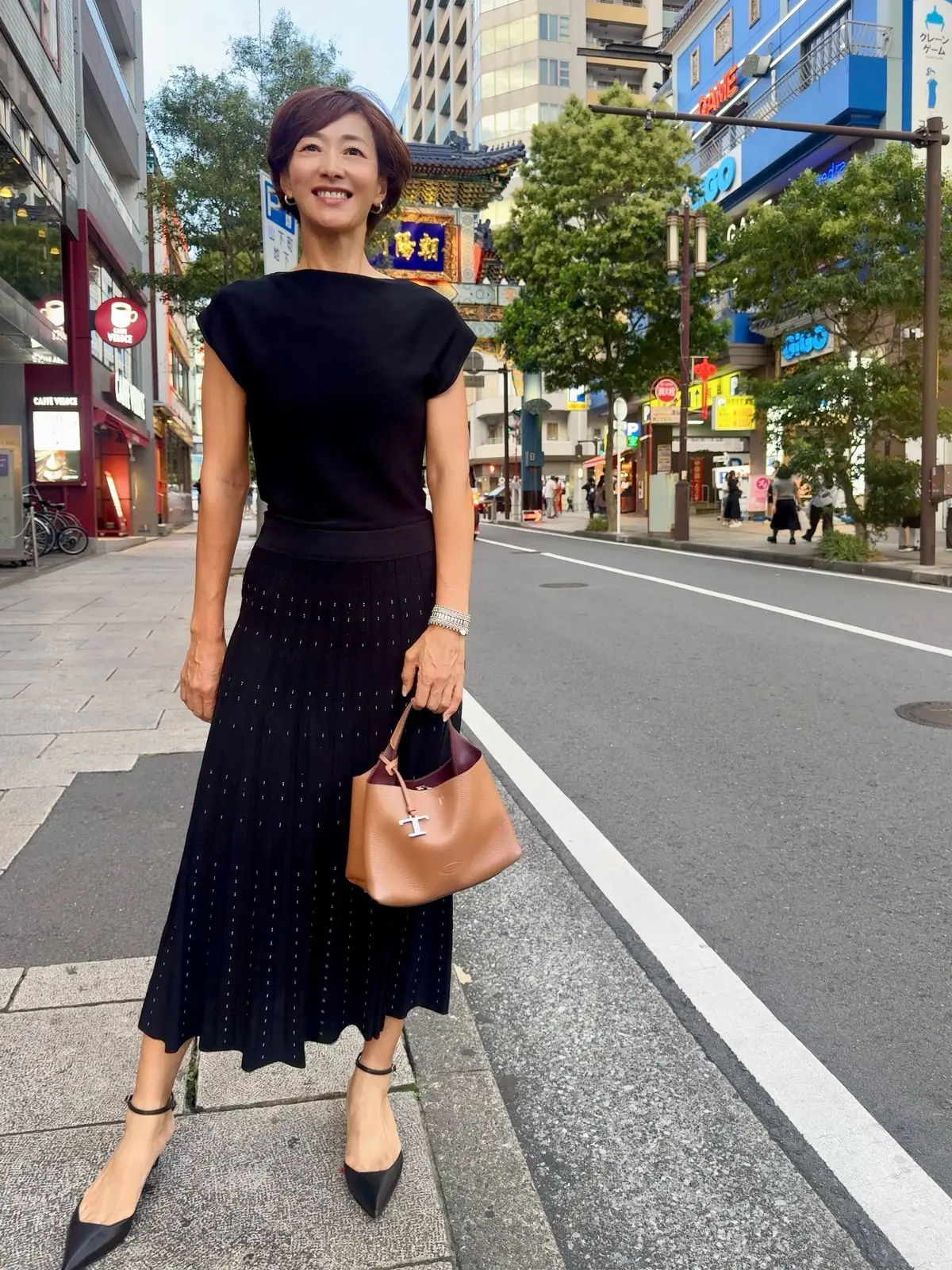 黒いトップスに黒のプリーツスカート、黒いヒールパンプスを着用した女性が、街並みを背景に立っている。右手にベージュのハンドバッグを持ち、笑顔を見せている。