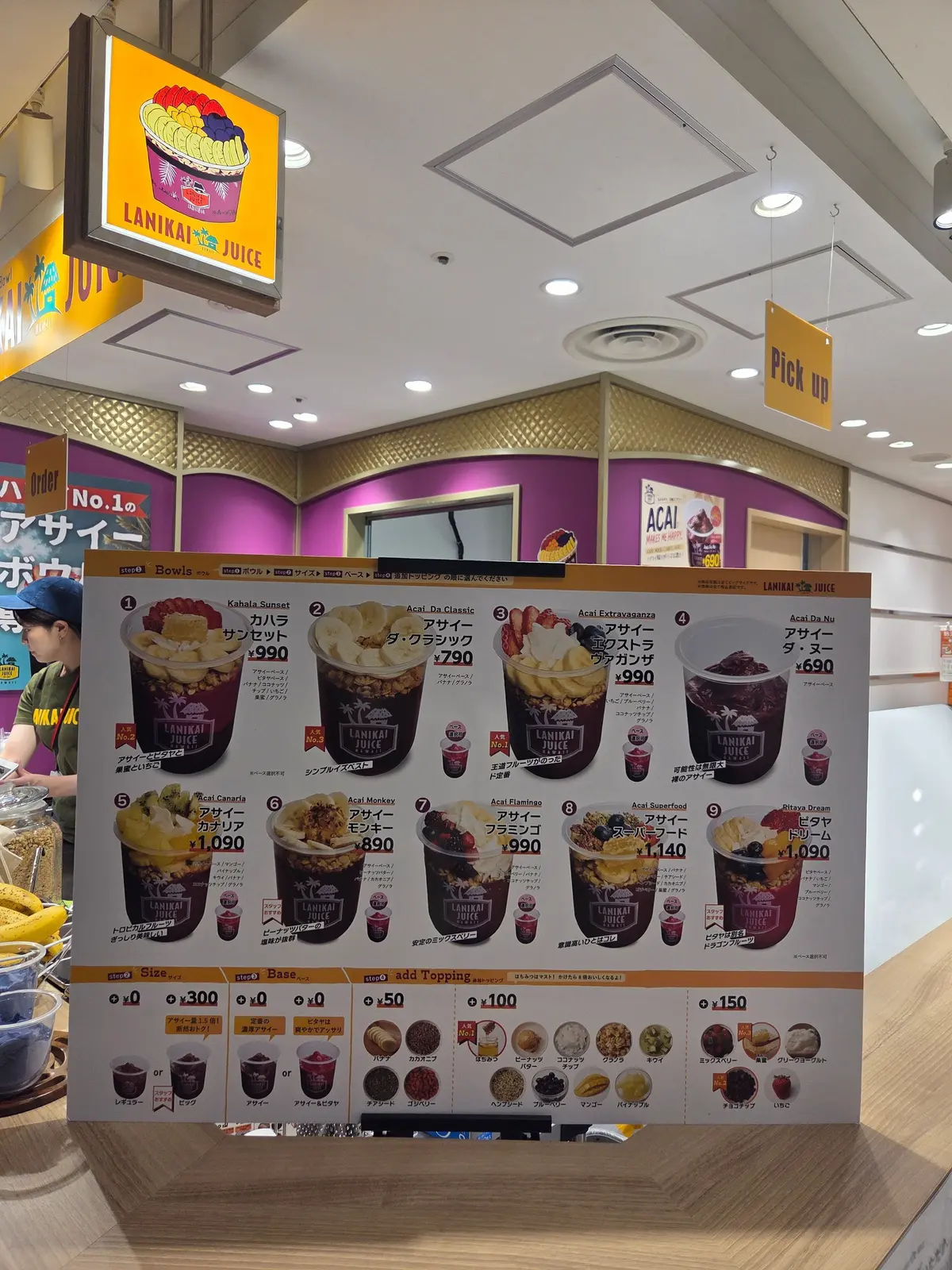 「LANI KAI JUICE」のメニューボード。アサイーボウルの写真とメニューが並び、サイズ、ベース、トッピングの選択肢が示されている。天井にはブランドロゴの照明が設置されている。