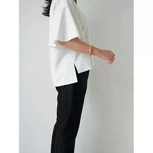 白いゆったりとしたTシャツと黒いパンツを着用した女性が、横向きに立っている写真。Tシャツの裾が片側だけ切りっぱなしになっている。右腕に金色のバングルを着用。背景は白。