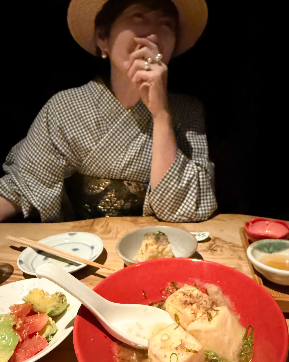 帽子を被った女性が、テーブルに並んだ日本料理を美味しそうに食べている様子。赤や白の小皿に盛り付けられた料理が並び、女性は白いシャツを着ている。
