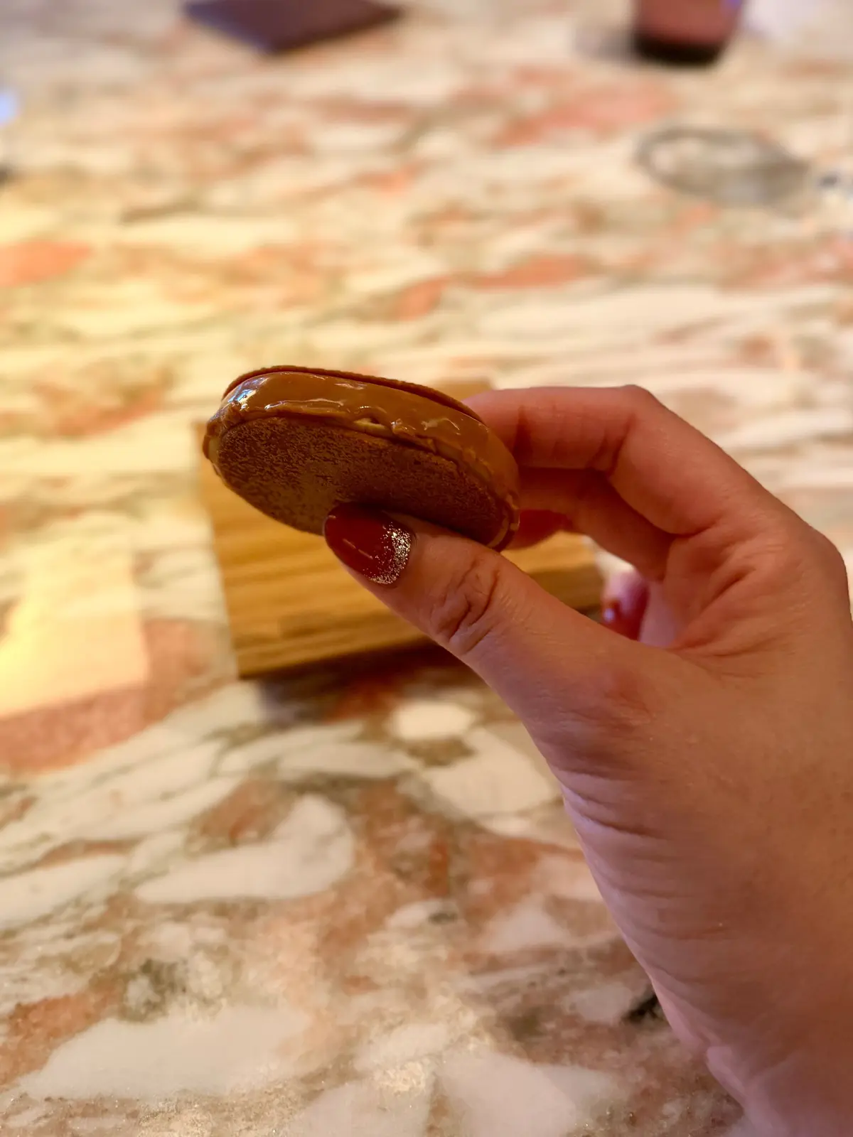 手はチョコレートコーティングされたサンドイッチクッキーを持ち、表面にはなめらかなチョコレートが広がっている。背景には木製のボードと石のテーブルトップが見える。