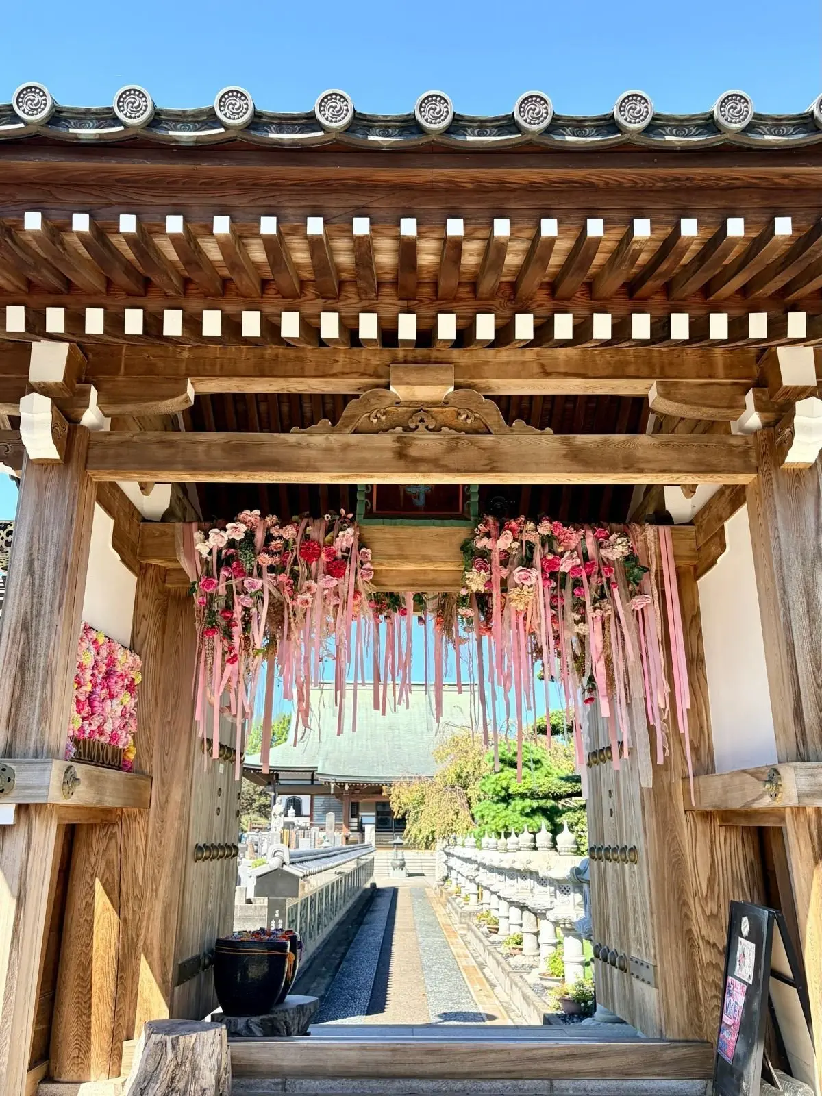 木造の鳥居にピンク色の花とリボンが飾られ、その先に石段と建物が見える。青空が背景に広がり、鮮やかな色彩が印象的。