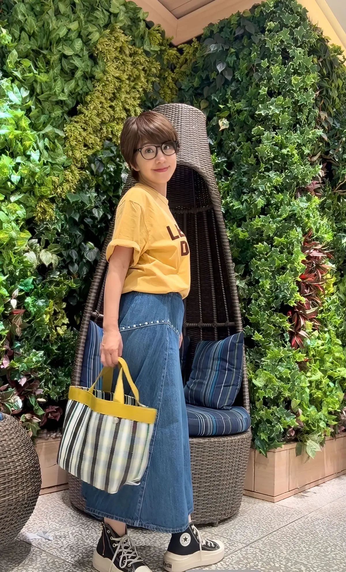 黄色のTシャツとデニージーンズを着た女性が、緑豊かな壁の前でポーズしています。彼女はストライプ模様のトートバッグを持ち、ワイドなスニーカーを履いています。背景には籐製のハンモックチェアがあります。