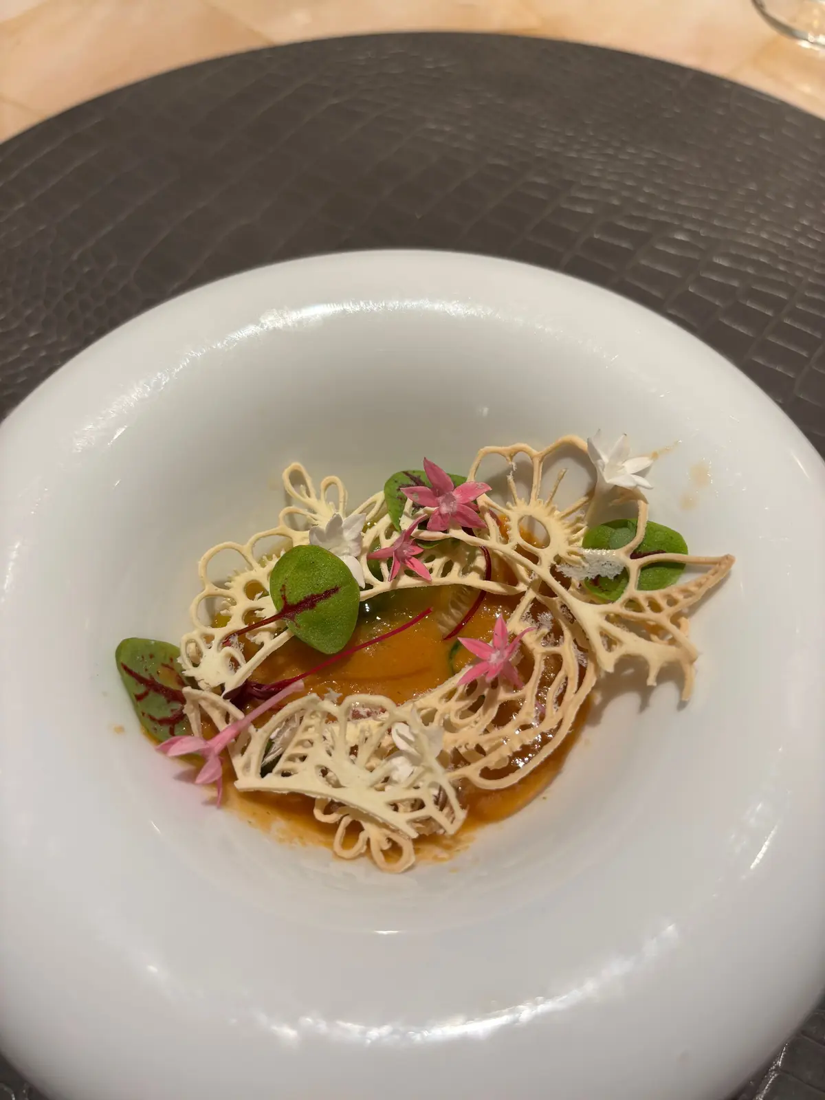 白いプレートに、細い麺のような具材と緑の葉、小さなピンクの花が飾られた料理。オレンジ色のソースが中心に広がっている。