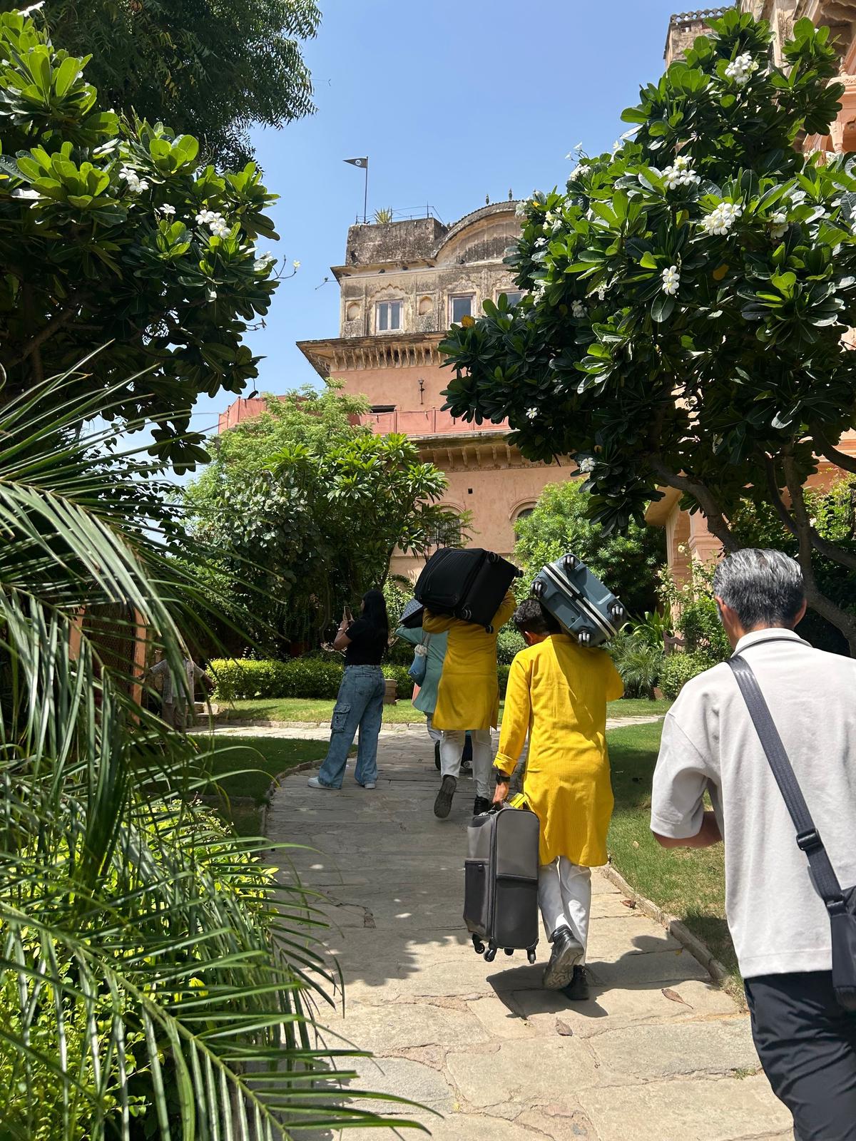 インドの庭園を歩く人々。黄色い服を着た人物がスーツケースを引いており、背景にはベージュ色の建物がある。緑豊かな木々が両側に茂り、白い花が咲いている。