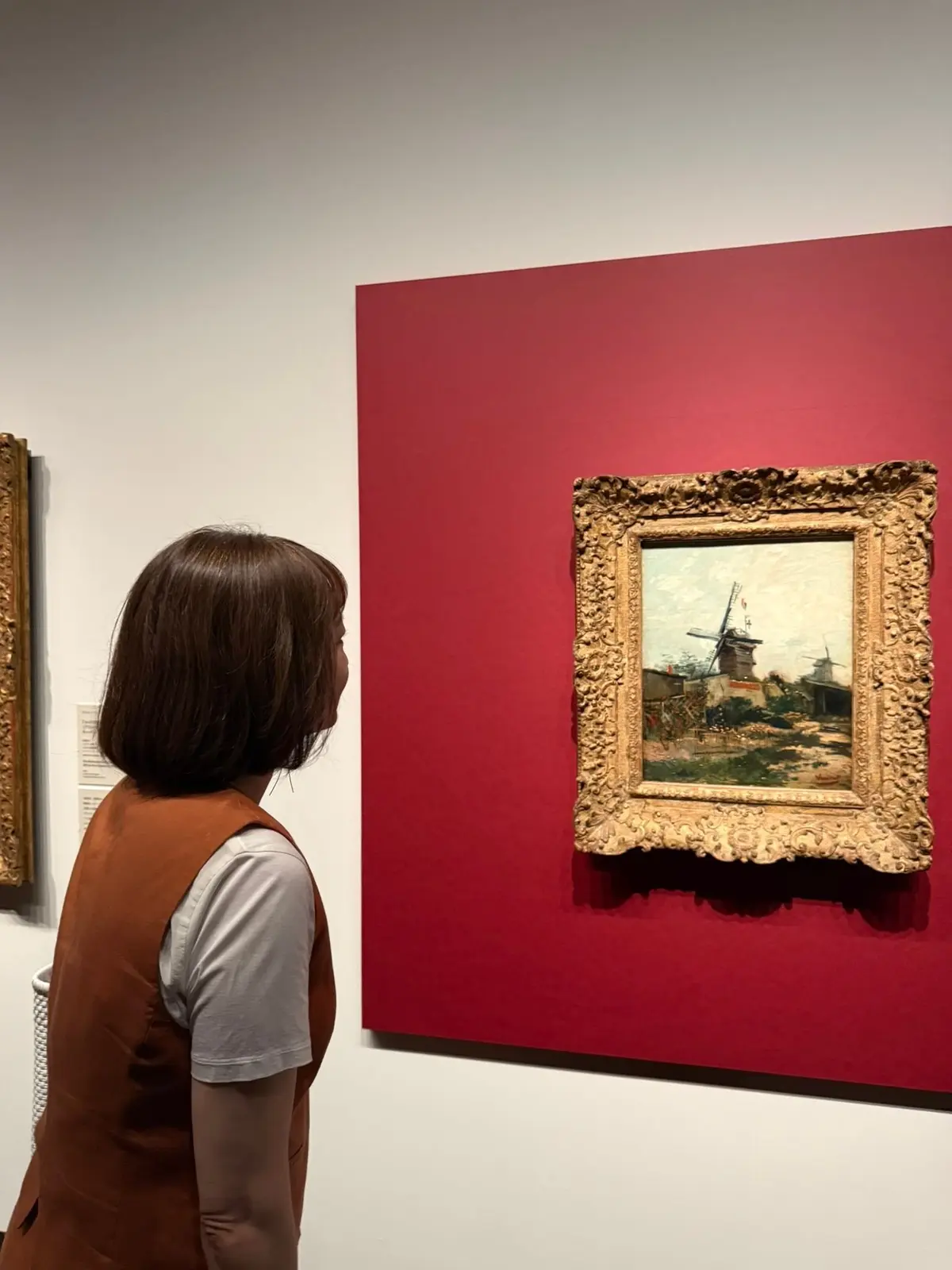 美術館で、女性が風車が描かれた絵画を鑑賞している様子。絵画は豪華な金色の額縁に入れられ、深紅の壁に飾られている。女性は茶色のベストを着用。