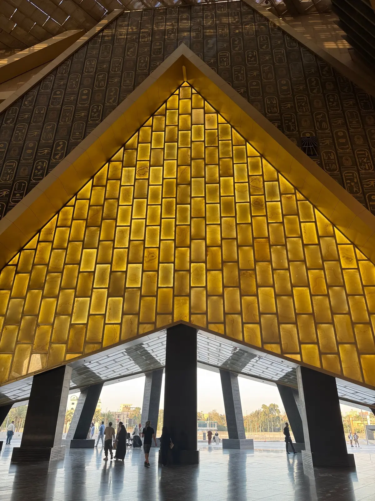 エジプト国立博物館の広大なロビー。金色の光が差し込むピラミッド型の壁面が特徴的で、上部には古代エジプトの象形文字が描かれている。黒い柱が並び、外の景色が見える開放的な空間。