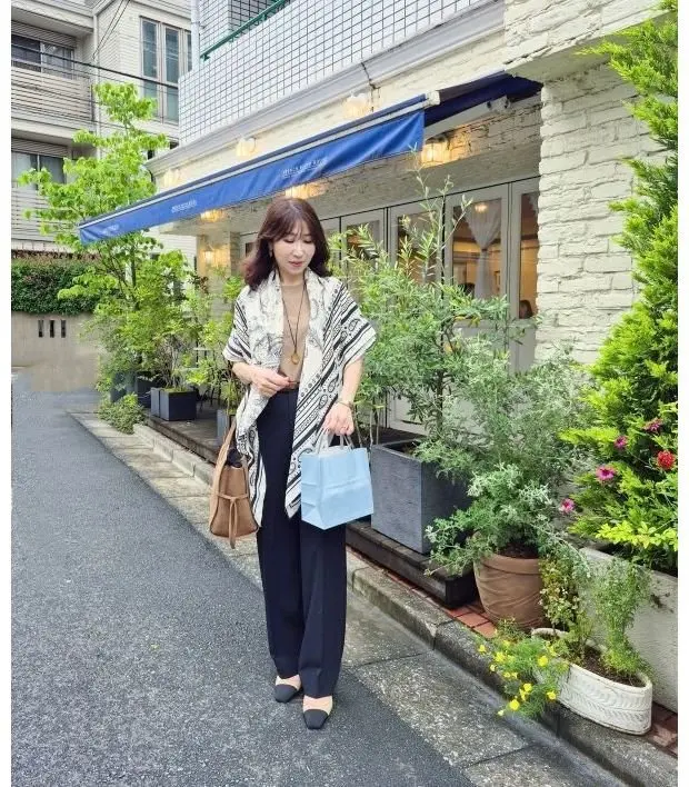 女性が青いカバノを手に、通り沿いの店前にてポーズ。白と黒のストライプカーディガン、黒のワイドパンツを着用し、ショルダーバッグを肩にかけ、明るい日差しの中、緑豊かな植え込みと建物背景が印象的。