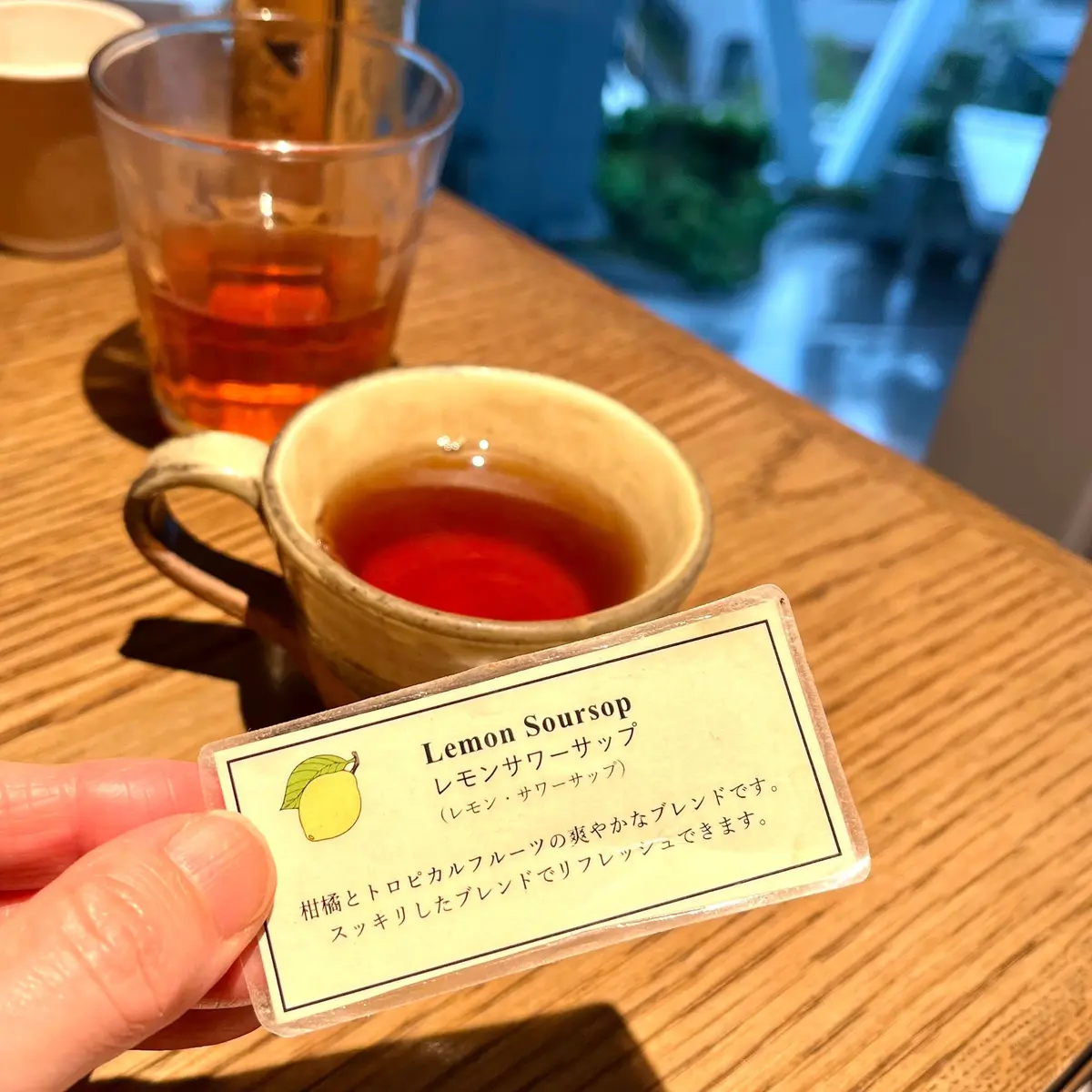 木のテーブルに茶色のグラスと白いマグカップにレモンの香りがする飲み物があり、指で持ったレモンソースの説明カードが置かれています。