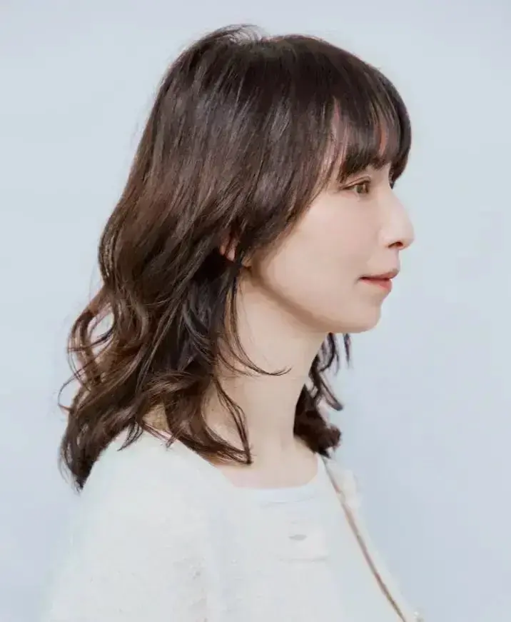 茶色の波状の髪が特徴の女性が、左を向いて淡い背景に立っている
