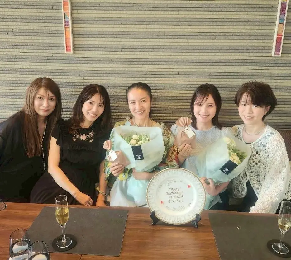 5人の女性が並んで座り、花束とシャンパンを持った誕生日のお祝い。中央に「Happy Birthday」のプレートが置かれている。
