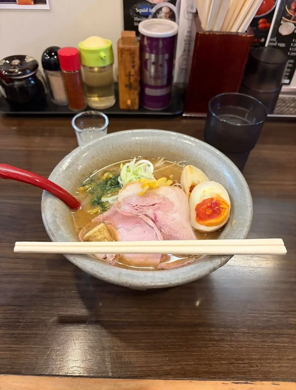 茶色のテーブルに置かれたラーメン。白地に灰色の模様が入った丼に、スープ、麺、ネギ、チャーシュー、半熟卵が入っている。赤い箸が置かれ、右にはグラスと調味料が置かれている。
