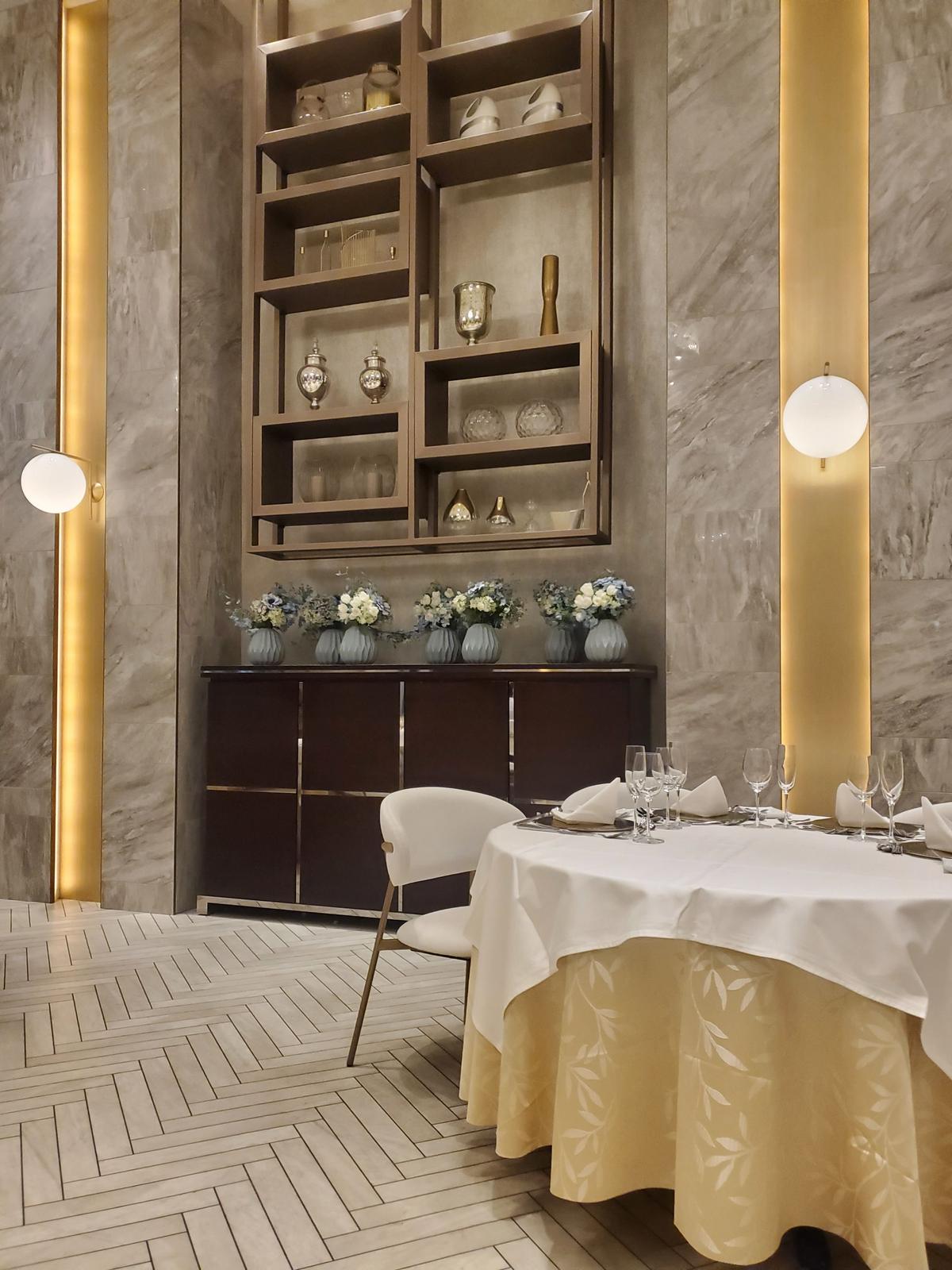 上品な内装のレストランの一角。丸テーブルに白いテーブルクロスが敷かれ、グラスやカトラリーが並んでいる。背後には、ワイングラスや花瓶が飾られた棚と、それに合わせた花束が置かれたキャビネットがある。壁はグレーの石材調で、照明が間接的に照らしている。