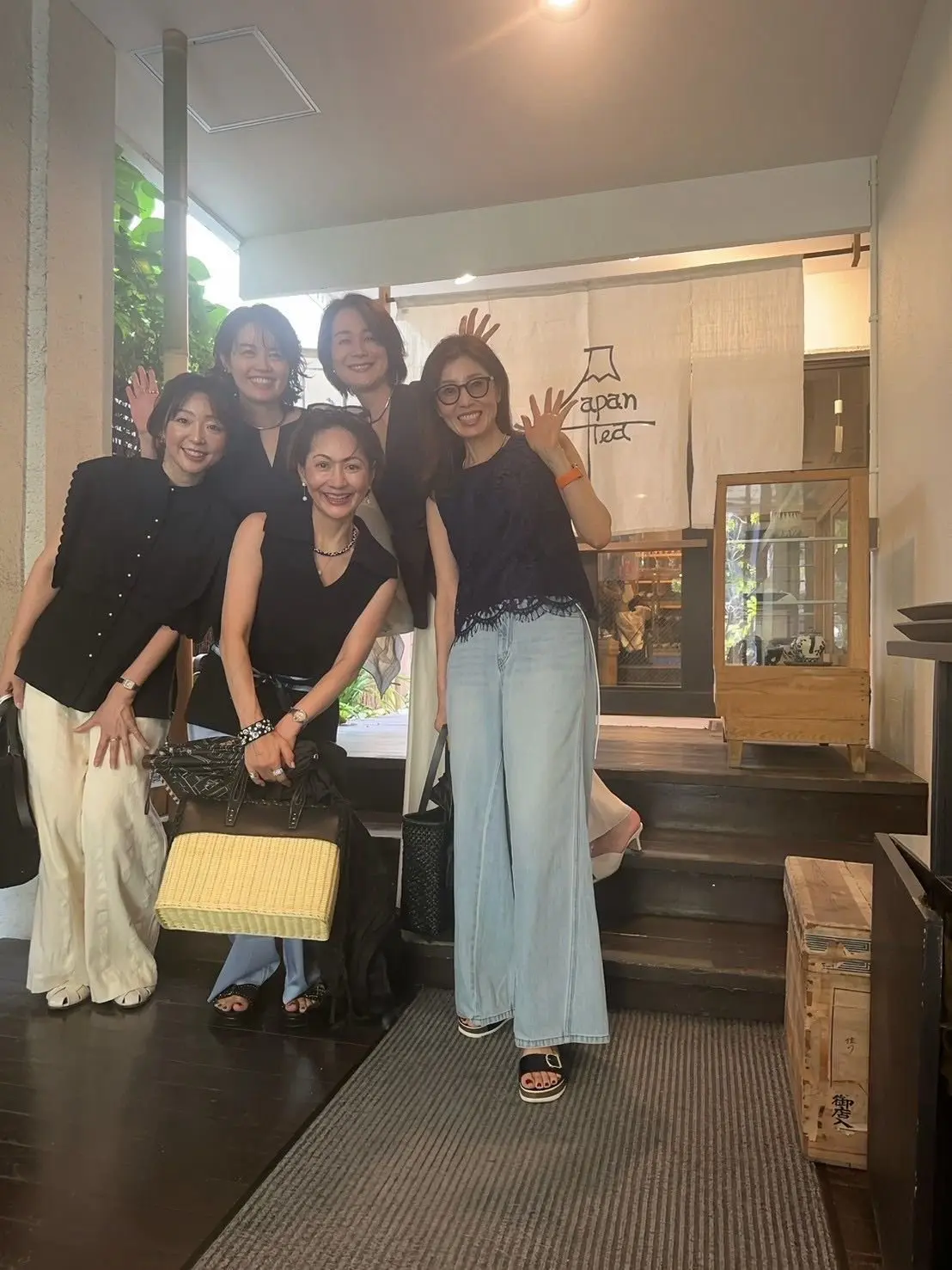 5人の女性が、白い壁の前に集まって笑顔でポーズをとっている。左から、黒の服を着た女性、白いパンツスタイル、長髪の女性、黒のトップスと黒いスカート、黒のトップスにデニムの女性、黒のワンピースを着た女性。
