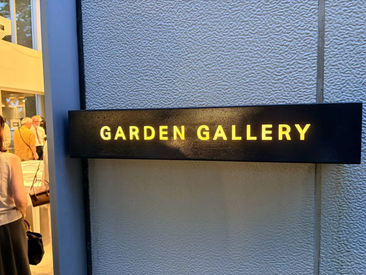 黒い長方形の看板に、黄色の文字で「GARDEN GALLERY」と表示されている。看板は壁に取り付けられており、背景には粗い質感の白い壁が見える。看板の左側には、窓から人物がぼんやりと見える。