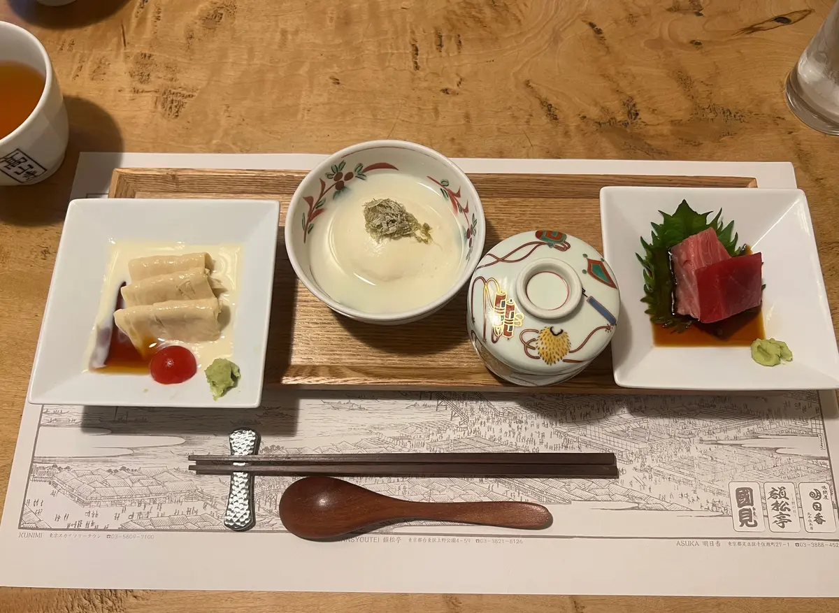 木製トレイに、四角い白い皿に蒸し魚の盛り合わせ、四角い白い器にワサビとマグロの刺身、小さな白い器に白いスープ、小さな丸い器に茶色いお茶、木製の箸とスプーンが並ぶ。