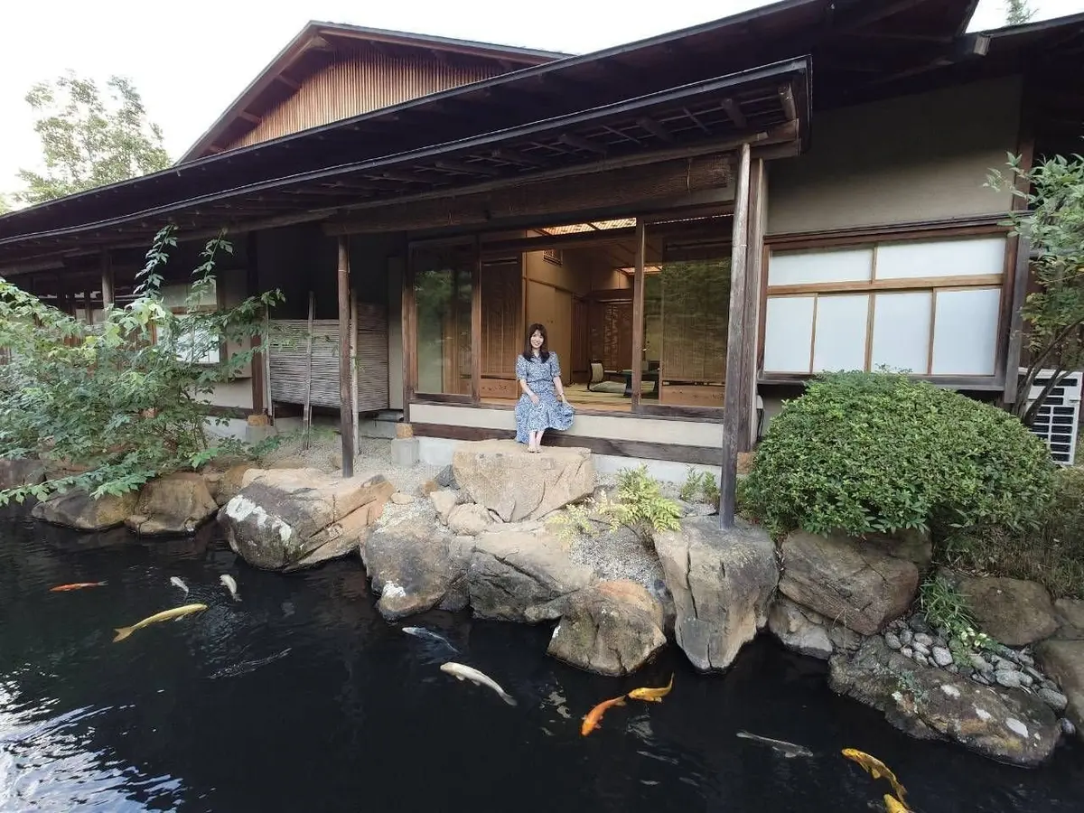 伝統的な日本の建物と庭園。女性が岩の上に立ち、池の鯉を眺めている。常磐ホテルの離れ「九重」。