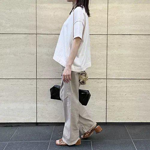 ベージュの壁に寄りかかった女性が、ベージュのワイドパンツと白の半袖シャツを着て、黒のクラッチとショルダーバッグを持ち、茶色のサンダルを履いている