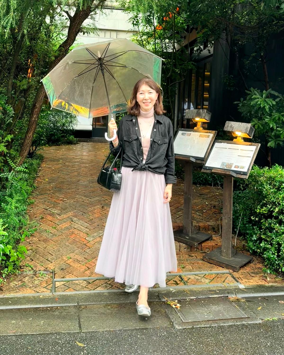 雨の中、女性が透明な傘をさし、笑顔でこちらを見ている。黒いタートルネックにデニムジャケット、チュールスカート、シルバーの靴を着用。黒いショルダーバッグを斜めにかけ、背景には緑豊かな植栽とレストランの看板がある。