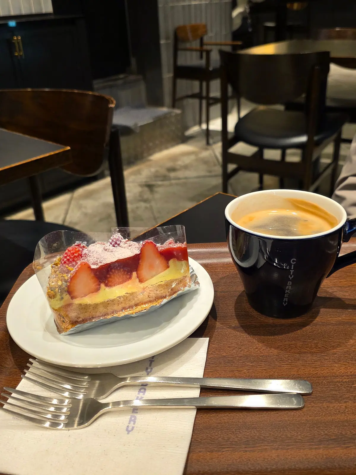 カフェのテーブルに置かれた、イチゴとクリームが乗ったパフェとコーヒー。フォークとナイフが、白いナプキンの上に置かれている。背景には、椅子とテーブルが見える。