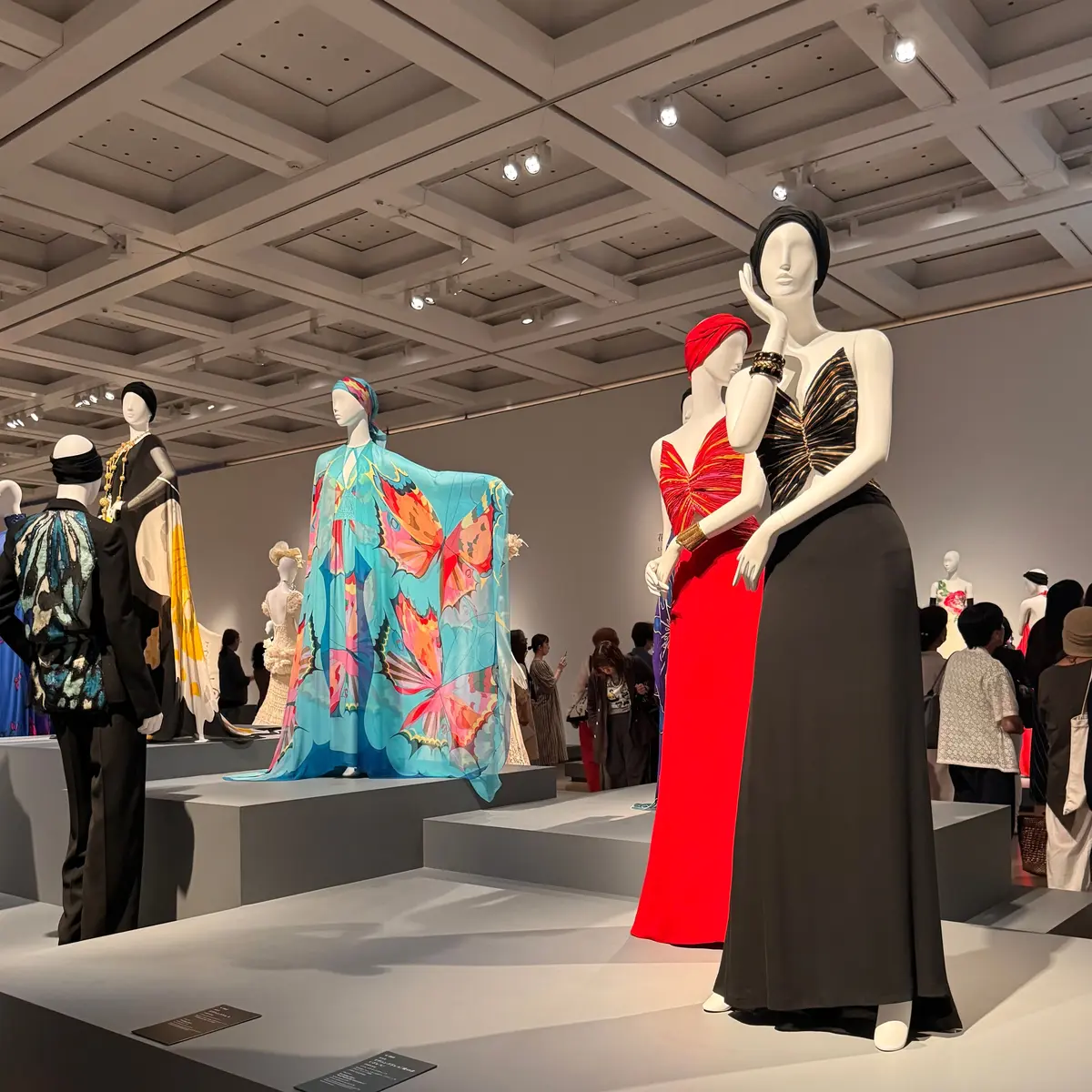 美術館の展示場で、複数のマネキンが様々なデザインのドレスを身につけている。鮮やかな水色のプリント、赤いシンプルなドレス、黒のロングドレスなど、多様な色彩とスタイルが並んでいる。背景には来場者が見られ、天井は格子状の照明が設置されている。