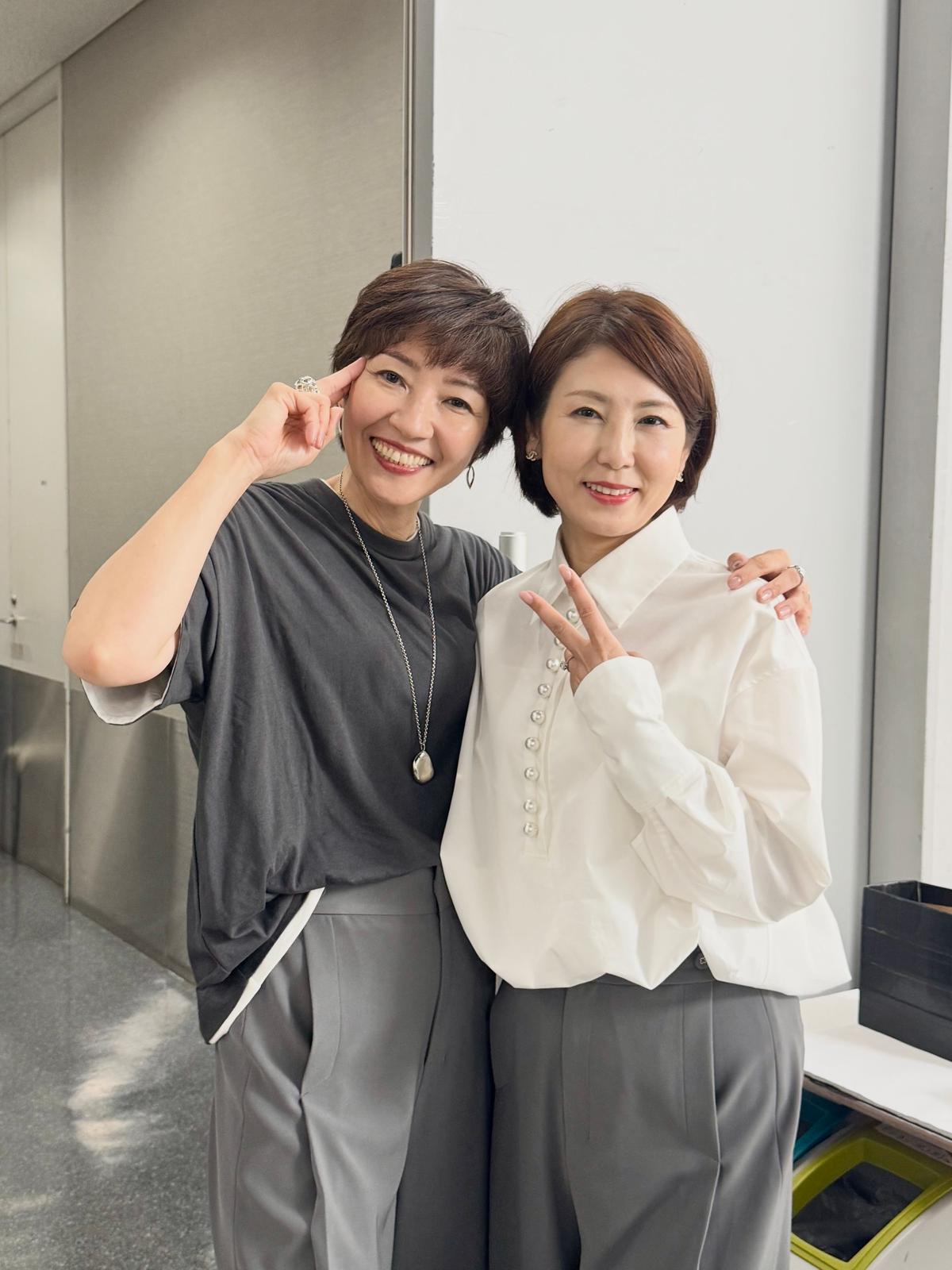 笑顔の2人の女性が並んで立っている写真。左の女性は黒いトップスとパンツを着用、右の女性は白いブラウスを着ている。背景は白い廊下で、右にはゴミ箱とテーブルがある。