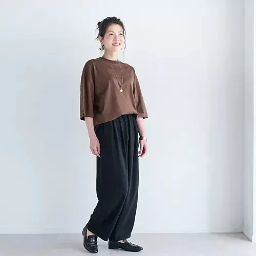 茶色のトップスと黒いワイドパンツを着用した女性が立っている全身写真。女性は右手をポケットに入れ、微笑んでいる。金色のネックレスと腕時計を着用。背景は白。
