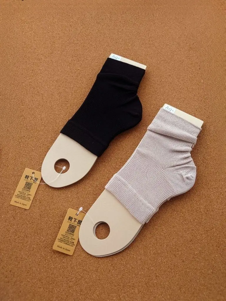 Ag+ブランドの黒と灰色の短い靴下2足が、茶色の背景に立てかけられている写真。靴下には白い縁取りがあり、それぞれにタグが付いている。
