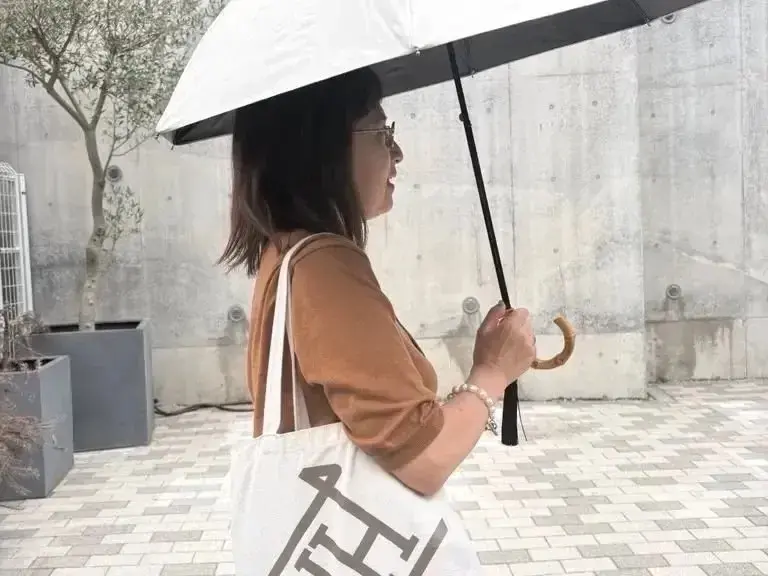 女性が白い傘を差して歩き、茶色の服を着て白いトートバッグを肩にかけた姿