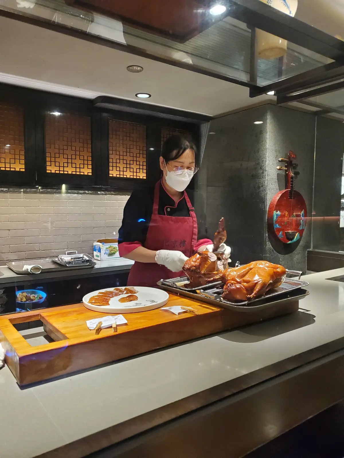 調理員がマスクと手袋を着用して、木製のカウンターに置かれた大きな焼き鳥をトレイに載せている。カウンターには切り身の肉と調味料が並んでいる。